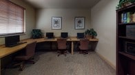 This single-floor business center is thoughtfully designed to support productivity and connectivity. The room features an L-shaped, light wood workstation along one wall, accommodating multiple desktop computers, keyboards, and mice, paired with comfortable upholstered rolling chairs. A second straight desk along an adjacent wall provides additional workspace, also equipped with computer stations. A window with horizontal blinds allows for controlled natural light in this section of the room.

On the opposite side of the space, two large, dark wood bookcases stand prominently, filled with an assortment of books and accented with decorative globe figurines and potted greenery on top. A recessed ceiling light illuminates this area, which also includes a standalone computer desk positioned near a glazed interior door. The multi-pane glass door offers a view into the adjacent corridor and allows light to filter through, contributing to an open feel.

The flooring throughout is carpeted in a neutral, patterned style that complements the warm beige tones of the walls. Framed botanical prints and lush indoor plants are placed throughout, adding a welcoming, professional ambiance. The layout is efficient and well-organized, making this space well-suited as a shared business or resource center.