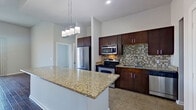 This single-floor residence offers a thoughtfully designed layout encompassing two bedrooms, two bathrooms, a combined kitchen and living area, a dedicated laundry room, and two closets. The open-concept kitchen is a standout feature, showcasing rich dark-brown cabinetry, granite countertops, a large center island with pendant lighting overhead, and a full suite of stainless steel appliances including a refrigerator, range, microwave, and dishwasher. A mosaic tile backsplash adds visual texture to the space. Tile flooring runs throughout the kitchen area, transitioning to warm hardwood in the living room, which benefits from tall ceilings, a ceiling fan, and sliding glass doors that invite natural light and provide outdoor access. The primary bedroom features hardwood flooring, high ceilings, a ceiling fan, and a sliding glass door to the exterior. The secondary bedroom is carpeted and receives natural light through a large window. A walk-in closet offers dual hanging rods for organized storage. The bathrooms include a full tub-shower with floor-to-ceiling tile surround. The laundry room provides wire shelving and dedicated washer and dryer hookups.
