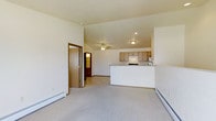 This single-floor space features an open-concept layout that connects the living area and kitchen, creating a spacious and functional floor plan. The living area is finished with light-toned wall-to-wall carpeting and neutral cream-colored walls, contributing to a bright and clean aesthetic. Vaulted ceilings add a sense of height and openness to the main living space, while a ceiling fan provides both air circulation and a subtle decorative element.

The kitchen is positioned toward the rear of the open area and features light wood cabinetry that provides ample storage. A white peninsula with a raised breakfast bar serves as a natural divider between the kitchen and living space, offering additional functionality for casual dining or meal prep. Recessed and flush-mount lighting fixtures illuminate the kitchen area effectively.

Baseboard heating units run along the lower walls, indicating a practical and efficient heating system suited for colder climates. A doorway visible on the left side of the living area leads to an additional room, suggesting further private living space beyond the open-plan area. Overall, the space maintains a clean, straightforward design with practical finishes throughout.