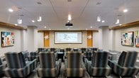 Located at 4451 Telfair Blvd in Camp Springs, Maryland, this single-floor venue is designed as a dedicated screening and presentation space. The room is thoughtfully arranged with rows of generously sized, upholstered lounge chairs in a muted teal and gray palette, offering comfortable seating for a sizeable audience. The layout provides clear sightlines to a large, wall-mounted projection screen framed by warm amber accent panels, which add a touch of visual warmth to the otherwise neutral interior. A ceiling-mounted projector is centrally positioned to deliver sharp, high-quality projection across the full screen surface. The ceiling features a recessed tile grid with integrated downlighting supplemented by elegant pendant lights, creating a well-layered and adjustable lighting environment suited for both presentations and screenings. Decorative wall sconces flank the projection wall, adding a refined finishing touch. Patterned carpeting underfoot contributes to the room's acoustically considered design, while the overall color scheme of warm whites, amber wood tones, and cool upholstery creates a professional yet inviting atmosphere. This space is well-suited for corporate presentations, training sessions, film screenings, or community events.