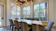 Located in Dearborn, MI, this thoughtfully designed single-floor communal space combines modern aesthetics with warm, inviting details throughout. The main gathering area is defined by soaring ceilings and floor-to-ceiling windows that flood the room with natural light and offer expansive views of the surrounding greenery. A striking multi-bulb chandelier hangs above a large rectangular dining table surrounded by bold black-and-white striped upholstered chairs, set atop a decorative area rug. A comfortable lounge seating area with tufted sofas and accent pillows is positioned along the window wall, creating a relaxed, social atmosphere. The adjacent lounge entry features shiplap accent walls, exposed dark wood ceiling beams, herringbone-patterned wood-look flooring, and eclectic checkered lounge chairs arranged around a live-edge coffee table. Art prints and a globe pendant chandelier add refined character to the hallway connecting the spaces. The fully equipped kitchen is a standout feature, showcasing bold black-and-white patterned encaustic tile backsplashes, white cabinetry with gold hardware, quartz countertops, stainless steel appliances, and open wood shelving. Herringbone tile flooring ties the kitchen's design together, making it both functional and visually distinctive. This space is ideal for gatherings, events, and everyday community use.