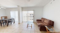 This single-floor, 2-bedroom, 1-bathroom residence offers a practical and comfortable layout designed to maximize everyday living. The living room is bright and airy, featuring light tile flooring, a textured ceiling, a wall-mounted air conditioning unit, and sliding glass doors fitted with vertical blinds that open to a balcony, allowing natural light to fill the space. Adjacent to the living area, the dining room continues the tile flooring and includes a ceiling fan, with additional access to outdoor views through another set of vertical blind-covered sliding doors. The kitchen is efficiently designed with warm wood cabinetry, granite-style countertops, a mosaic tile backsplash, and a full suite of stainless steel appliances including a refrigerator, electric range, over-the-range microwave, and dishwasher. Both bedrooms are carpeted and offer a quiet, restful atmosphere; each includes a built-in closet with louvered doors and wall-mounted air conditioning units for individual climate control. The shared bathroom is finished with tile flooring, a wood vanity topped with granite countertops, a large mirror, and a full shower-tub combo. A connecting hallway ties the layout together, providing smooth flow throughout the home.