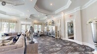 This single-story clubhouse in Durham, NC, offers a generously sized, thoughtfully designed communal space that blends sophistication with comfort. The entrance foyer makes an immediate impression with polished marble-style tile flooring, a tiered crystal chandelier, and a tray ceiling framed by refined crown molding. Large glass-paneled doors flood the space with natural light and provide views of the surrounding community.

The main lounge area features an octagonal tray ceiling, a white marble fireplace flanked by large windows with decorative valances, and plush seating arranged around curated sculptural décor. Patterned carpeting adds warmth and texture throughout this central gathering area.

The kitchen is well-appointed with rich walnut-toned cabinetry, white quartz countertops, a gleaming marble-style tile floor, and a central island with bar seating—ideal for events and casual gatherings. An adjacent dining area provides multiple seating options, including high-top bistro tables and a formal dining set, all bathed in natural light from oversized windows overlooking the community exterior.

Throughout, the clubhouse maintains a cohesive, upscale aesthetic defined by neutral tones, elegant architectural details, and carefully selected décor.