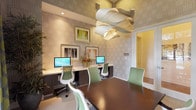 This single-floor business center is thoughtfully designed to support both focused work and group collaboration. The space is divided into two functional zones: a computer workstation area and a conference area. The workstation zone features a built-in counter with dark cabinetry below, accommodating multiple desktop computers and ergonomic green-and-white chairs. Framed artwork and fresh floral arrangements add warmth to this area.

The conference zone centers around a rich, dark walnut table surrounded by matching green-and-white ergonomic chairs. A striking sculptural pendant light with layered, ribbon-like forms serves as the focal point overhead, drawing the eye upward toward the tall ceilings. The walls throughout are covered in a sophisticated geometric patterned wallcovering in soft neutral tones.

Large black-framed windows flood the conference area with natural light and offer views of lush greenery outside, complemented by green-and-yellow patterned drapes. A bold landscape-inspired canvas artwork anchors one wall, adding a vibrant visual contrast. French doors with glass panels connect the space to an adjacent area, enhancing openness. The neutral carpet flooring, curated artwork, and cohesive color palette create a polished, professional ambiance.