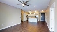 This single-floor space showcases an open-concept layout that seamlessly connects the living area to the kitchen, creating a spacious and airy atmosphere. The rich, dark hardwood flooring runs throughout, adding warmth and a sense of continuity across the entire space.

The kitchen is a standout feature, designed with crisp white cabinetry that extends to the upper walls, providing ample storage. A central island with a dark countertop serves as both a functional workspace and a natural gathering point. Stainless steel appliances, including a refrigerator and range, complement the modern aesthetic, while a built-in microwave adds convenience. The sink is positioned on the island, enhancing workflow within the kitchen.

Lighting is thoughtfully layered throughout the space. Pendant lights hang above the kitchen island, recessed lighting is positioned strategically across the ceiling, and a ceiling fan with an integrated light fixture anchors the living area. The neutral, warm-toned walls provide a versatile backdrop that complements both the white cabinetry and dark flooring. The entry door, finished in a deep charcoal, adds a bold accent element to the overall design.