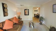 This single-floor residence offers a welcoming and cohesive layout, with the living room serving as the central hub of the space. The living area features plush, earth-toned seating accented by vibrant orange throw pillows, creating a warm and inviting atmosphere. Crown molding lines the ceiling throughout, adding a refined architectural touch, while neutral-toned carpeting grounds the space comfortably underfoot.

A decorative framed mirror adorns the living room wall, enhancing the sense of depth and light, while a ceramic table lamp on a side table provides soft, ambient illumination. A glass-top coffee table adds a contemporary accent to the traditionally styled room.

Through an elegant arched doorway, the dining area is seamlessly connected, featuring hardwood-style flooring that contrasts with the carpeted living space. A ceiling fan with a light fixture keeps the dining area bright and comfortable. The dining set, framed by a decorative wall piece, provides a functional yet stylish space for meals.

A lush potted plant near the entrance adds a natural element, while the overall color palette of warm beiges and soft whites lends the home a calm, approachable ambiance throughout.