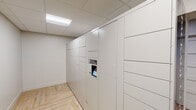 This single-floor amenity space is thoughtfully designed to offer both convenience and wellness facilities. The package room is the standout functional feature, lined wall-to-wall with a Package Concierge automated locker system. The lockers are finished in a clean, neutral white, complementing the bright drop-tile ceiling with recessed fluorescent lighting. The floor throughout features light wood-toned herringbone-patterned vinyl plank, adding a warm and modern aesthetic to the utilitarian space. The room is neatly proportioned, offering ample locker capacity in a range of sizes to accommodate deliveries of varying dimensions. A glass-paneled door separates the package area from an adjacent corridor, where a fire extinguisher and access panel are mounted, and natural light hints at an outdoor area beyond.

The fitness center, accessible via a full-length glass door, is well-appointed with a selection of free weights including a full dumbbell rack, cardio and strength-training equipment, and a wall-mounted flat-screen television. Patterned carpet tiles in tones of blue, green, and gold cover the gym floor, and an arched window allows natural light to fill the space. Security and utility panels are also present within the building, underscoring the functional infrastructure supporting the overall facility.