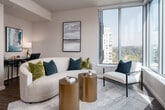 This thoughtfully designed single-floor residence showcases a seamless blend of contemporary style and functional living. The open-concept layout connects the kitchen, dining, and living areas, creating a cohesive and inviting atmosphere. The kitchen is a standout feature, anchored by a large island with striking marble-veined quartz countertops, dark wood cabinetry, and stainless steel appliances including a refrigerator, microwave, and range. A statement pendant light in brushed gold hangs above the island, complementing the recessed lighting overhead. Wood-look flooring runs throughout the main living areas, adding warmth and continuity. The living space benefits from large windows that invite abundant natural light and offer views of the surrounding urban environment. A textured concrete column adds an architectural accent, lending an industrial-chic character to the open plan. The bedroom is a serene retreat, featuring a wide window with city views, neutral tones, and warm ambient lighting. The bathroom is well-equipped with a soaking tub, large-format tile surround, a floating vanity with navy cabinetry, and an illuminated mirror that provides both style and practicality. Overall, the residence exudes a polished, modern aesthetic with high-quality finishes and a cohesive design language throughout.