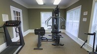 This single-floor fitness room offers a dedicated workout space designed for strength training and exercise. The room is finished with olive-green painted walls accented by white crown molding and white baseboards, lending a clean, polished appearance. Carpeted flooring in a neutral tone provides comfort underfoot throughout the space. Natural light enters generously through multiple windows fitted with horizontal blinds, creating a bright and welcoming environment. The centerpiece of the room is a multi-station cable and weight machine, suitable for a variety of upper and lower body exercises, including lat pulldowns, chest presses, and leg extensions. The equipment is positioned to allow adequate clearance for safe use. Overhead fluorescent lighting supplements natural light, ensuring the space remains well-lit at all times. The overall layout is straightforward and functional, with the fitness equipment arranged to maximize the usable floor area. The room's simple, uncluttered design keeps the focus on utility, making it a practical space for residents or users seeking convenient access to strength training equipment without the need to travel to an off-site facility.