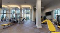 This well-appointed fitness center is designed to accommodate a comprehensive range of workout activities within a single, expansive open-plan floor. The space is characterized by soaring ceilings that lend an airy, open feel throughout, complemented by large floor-to-ceiling windows that frame views of lush outdoor greenery and palm trees, flooding the interior with natural light.

The layout is thoughtfully organized into distinct workout zones. A dedicated cardio area houses a row of elliptical trainers, stationary bikes, treadmills, and cycling bikes, positioned to take advantage of the natural light from the oversized windows. A strength training section features a full rack of dumbbells displayed along a large wall-mounted mirror, flat and adjustable benches, and a variety of cable and resistance machines, all finished in a bold yellow and grey color scheme.

A functional training zone offers medicine ball racks, stability balls, and open floor space for dynamic exercises. The flooring throughout consists of a durable checkered carpet tile in neutral grey and tan tones, providing both comfort and practicality. Linear LED lighting fixtures are suspended from the ceiling, ensuring bright, even illumination. Structural columns are integrated seamlessly into the design, and large mirrors are strategically placed to enhance the sense of space.