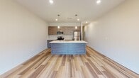This contemporary space presents a clean, modern aesthetic throughout, with a thoughtfully designed layout spread across multiple levels connected by a carpeted staircase. The open-concept main living area is anchored by a sleek kitchen featuring warm wood-tone cabinetry, white quartz countertops, and a full suite of stainless steel appliances including a range, microwave, and refrigerator. A central island with an integrated sink and pendant lighting above serves as both a functional workspace and a natural gathering point. Light-toned wood-style flooring flows throughout the main living areas, enhancing the bright, airy atmosphere.

The living space extends to a balcony accessible through sliding glass doors, allowing natural light to fill the room. A ceiling fan with recessed lighting overhead adds comfort and functionality. Bedrooms are finished with plush carpeting and ceiling fans, providing a quieter, softer environment. One carpeted room features a window, while another offers a more enclosed setting.

The bathroom is finished in a clean, minimalist style with light wood-tone cabinetry, a white countertop vanity, and a full tub-shower combination. A dedicated laundry area houses a full-sized side-by-side washer and dryer, adding everyday convenience to this well-appointed space.