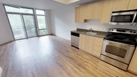 Located in Seattle's Delridge neighborhood, Unit 213 at 4040 26th Ave SW offers a thoughtfully designed single-floor layout with a clean, contemporary aesthetic. The open-concept living and kitchen area is anchored by warm-toned hardwood-style flooring that flows seamlessly throughout the unit, creating a cohesive and inviting atmosphere. Large floor-to-ceiling windows in the living area flood the space with natural light and provide a lush, green outlook, enhancing the sense of openness.

The kitchen is efficiently designed with light maple cabinetry, gray laminate countertops, and a full suite of stainless steel appliances, including an electric range, dishwasher, and over-the-range microwave. Ample upper and lower cabinet storage makes the kitchen both practical and visually streamlined.

The living area is generously proportioned, offering flexible space for a variety of furniture arrangements, with clean white walls that provide a neutral canvas. The bathroom features a light maple vanity with a warm-toned countertop, a large wall mirror, and a full-size soaking tub with a shower. Hardwood-style flooring continues into the bathroom, maintaining design consistency throughout the unit. Overall, the space presents a modern, well-appointed residence suited for comfortable urban living.