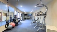 This fitness center offers a comprehensive workout environment designed to accommodate a variety of exercise routines and fitness goals. The space is laid out on a single floor, providing an open and accessible layout that allows for easy navigation between different workout zones.

The cardio area features multiple treadmills positioned toward the back of the room, with a wall-mounted flat-screen television providing entertainment during workouts. Natural light filters in through windows along one wall, complementing the recessed ceiling lighting to keep the space bright and energizing.

The free weights section is well-stocked with a full rack of dumbbells in varying sizes, along with medicine balls and kettlebells neatly arranged for easy access. An adjustable weight bench with a barbell rack provides a dedicated space for strength training exercises.

Functional training is supported by TRX suspension straps anchored to the ceiling, along with stability balls and additional resistance tools. Mirrored wall panels in the strength training area allow users to monitor their form during exercises.

The flooring throughout the space combines cushioned carpet and rubberized matting in key workout zones, offering both comfort and safety. The neutral-toned walls and clean, organized layout contribute to a focused and motivating atmosphere suitable for fitness enthusiasts of all levels.