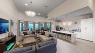 This single-floor community clubhouse is designed as an open, multi-functional gathering space that seamlessly blends comfort and functionality. Soaring ceilings in the main lounge area create a grand, airy atmosphere, accentuated by statement pendant lighting and large windows that flood the space with natural light and offer views of the surrounding landscape.

The centerpiece of the space is a well-appointed kitchen featuring white shaker cabinetry, stainless steel appliances, a striking marble-topped island with bar seating, and decorative wall sconces that add a touch of elegance. The kitchen transitions naturally into the open lounge area, which is furnished with plush neutral sofas and accent chairs adorned with navy blue cushions, centered on layered area rugs over light wood-style flooring.

Multiple dining areas are positioned near large windows dressed with layered curtains, providing serene outdoor views and ample natural light. Round wooden dining tables with upholstered chairs offer intimate seating arrangements throughout.

A dedicated media and business center lounge features a wall-mounted television, a built-in desk with multiple computer workstations illuminated by geometric pendant lights, and casual seating — ideal for both productivity and relaxation. Curated artwork and decorative accents throughout tie the space together with a cohesive, contemporary coastal aesthetic.