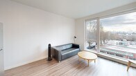 Located on Stone Way N in Seattle's vibrant Wallingford neighborhood, this one-bedroom, one-bathroom unit offers a thoughtfully designed, modern interior with a clean, minimalist aesthetic throughout. Light wood-tone flooring flows consistently across the entire space, complementing the bright white walls and recessed lighting that keep the unit feeling open and airy.

The combined kitchen and living area forms the heart of the unit. The kitchen is outfitted with stainless steel appliances — including a full-size refrigerator, electric range, built-in microwave, and dishwasher — paired with warm wood-grain cabinetry, white countertops, and upper white cabinets for ample storage. A floating wood shelf adds a functional design accent. The living area benefits from a large picture window that brings in generous natural light and offers views of the tree-lined street below.

A hallway connects the main living space to the private bedroom, a dedicated closet, and the combined bathroom and laundry room. The bathroom features a white undermount sink set in wood-grain cabinetry, a large mirror, and a shower. In-unit laundry adds everyday convenience. The entrance and hallway are clean and functional, with a utility panel neatly integrated into the corridor. The overall design prioritizes efficiency, comfort, and modern style.