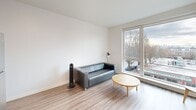 Located on Stone Way N in Seattle's vibrant Wallingford neighborhood, this one-bedroom, one-bathroom unit offers a thoughtfully designed, modern interior with a clean, minimalist aesthetic throughout. Light wood-tone flooring flows consistently across the entire space, complementing the bright white walls and recessed lighting that keep the unit feeling open and airy.

The combined kitchen and living area forms the heart of the unit. The kitchen is outfitted with stainless steel appliances — including a full-size refrigerator, electric range, built-in microwave, and dishwasher — paired with warm wood-grain cabinetry, white countertops, and upper white cabinets for ample storage. A floating wood shelf adds a functional design accent. The living area benefits from a large picture window that brings in generous natural light and offers views of the tree-lined street below.

A hallway connects the main living space to the private bedroom, a dedicated closet, and the combined bathroom and laundry room. The bathroom features a white undermount sink set in wood-grain cabinetry, a large mirror, and a shower. In-unit laundry adds everyday convenience. The entrance and hallway are clean and functional, with a utility panel neatly integrated into the corridor. The overall design prioritizes efficiency, comfort, and modern style.