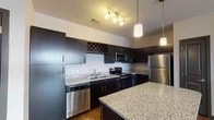 This single-floor residence at Springs At Hamburg offers a well-designed layout with a cohesive modern aesthetic throughout. The kitchen serves as a central highlight, featuring rich dark cabinetry paired with speckled granite countertops, a full suite of stainless steel appliances including a refrigerator, range, microwave, and dishwasher, and a built-in wine rack for added character. A granite-topped island provides additional prep space and casual seating, while pendant lighting and a track light fixture illuminate the area effectively. Wood-style plank flooring extends through the kitchen and into the open living area, which benefits from multiple large windows and a glass-paneled entry door that draw in generous natural light. A ceiling fan is centrally positioned in the living space, enhancing comfort. The bedrooms are carpeted for warmth and comfort, each featuring wide window banks with outdoor views and ceiling fans. The bathroom includes a full tub and shower combination with a curved curtain rod and wood-style flooring, maintaining a clean, functional aesthetic consistent with the rest of the unit.