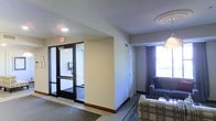 This single-floor space offers a straightforward and functional layout well-suited for extended stays or transitional living. The entryway is defined by a set of glass-paneled double doors with a classic black frame, complete with an illuminated exit sign overhead and a wall-mounted telephone — details that speak to the space's hospitality or institutional character. A carpeted foyer transitions smoothly into the main living area, where neutral beige walls and a textured ceiling create a clean, understated backdrop. The living area is furnished with a plaid-upholstered sofa and a dark leather chair, offering a cozy seating arrangement. A large window dressed with dark curtains allows natural light to filter into the room, brightening the space and providing a view to the exterior. Recessed and flush-mount ceiling fixtures provide consistent overhead lighting throughout. A closet door is visible along one wall, adding practical storage to the layout. The overall design is utilitarian yet comfortable, with warm neutral tones, durable flooring, and efficient use of space contributing to an environment that balances accessibility with everyday functionality.
