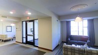 This single-floor space offers a straightforward and functional layout well-suited for extended stays or transitional living. The entryway is defined by a set of glass-paneled double doors with a classic black frame, complete with an illuminated exit sign overhead and a wall-mounted telephone — details that speak to the space's hospitality or institutional character. A carpeted foyer transitions smoothly into the main living area, where neutral beige walls and a textured ceiling create a clean, understated backdrop. The living area is furnished with a plaid-upholstered sofa and a dark leather chair, offering a cozy seating arrangement. A large window dressed with dark curtains allows natural light to filter into the room, brightening the space and providing a view to the exterior. Recessed and flush-mount ceiling fixtures provide consistent overhead lighting throughout. A closet door is visible along one wall, adding practical storage to the layout. The overall design is utilitarian yet comfortable, with warm neutral tones, durable flooring, and efficient use of space contributing to an environment that balances accessibility with everyday functionality.
