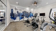 The Scout Landing Premier Apartments fitness center is a compact, well-appointed single-room facility designed to meet the everyday workout needs of its residents. The space is finished with neutral carpet tiles and bright white walls adorned with crown molding, contributing to a clean and polished aesthetic throughout.

A standout feature is the large, vivid mural spanning one full wall, depicting a stylized Nashville skyline in deep blues and whites alongside the Scout Landing branding — giving the room a distinct local identity. Beneath the mural sits a bank of personal storage lockers, offering convenient secure storage for residents during their workouts.

The cardio equipment lineup is well-rounded, including upright and recumbent stationary bikes, elliptical cross-trainers, and stair climbers from the True Fitness brand. Yoga and exercise balls, resistance bands, and foam rollers are neatly organized in a dedicated corner rack, supporting stretching and functional training. A wall-mounted flat-screen TV provides entertainment during workouts.

Natural light pours in through large windows fitted with blinds along one wall, while recessed ceiling lights and a ceiling fan ensure the room remains bright and comfortable at any time of day. A full-length glass partition wall opens the space visually to an adjacent lounge or common area, enhancing the sense of openness despite the room's intimate footprint.
