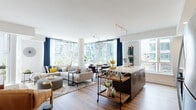 This thoughtfully designed single-floor residence combines contemporary style with functional living. The open-concept main living area is a standout, bathed in abundant natural light streaming through expansive floor-to-ceiling windows that frame views of the surrounding urban skyline. The living and dining areas flow seamlessly together, anchored by warm hardwood flooring and accented by a globe pendant light fixture overhead.

The kitchen is a study in modern design, featuring two-tone cabinetry with upper light grey and lower dark wood-finish cabinets, crisp white quartz countertops, a hexagonal penny-tile backsplash, stainless steel appliances, and a generous island that doubles as a casual dining surface.

A welcoming hallway connects the private areas of the home, lined with decorative wall art and louvered closet doors, leading to a well-appointed bedroom with large windows, hardwood floors, and generous storage. The bathroom is finished with large-format grey wall tiles, a soaking tub with shower, and clean white vanity surfaces.

A cozy sitting nook with a tufted leather chair, display cabinetry, and a marble-topped sideboard adds a refined lounge-like quality to the space. Throughout, the residence maintains a cohesive, modern urban aesthetic with warm tones and curated décor.
