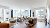 This thoughtfully designed single-floor residence combines contemporary style with functional living. The open-concept main living area is a standout, bathed in abundant natural light streaming through expansive floor-to-ceiling windows that frame views of the surrounding urban skyline. The living and dining areas flow seamlessly together, anchored by warm hardwood flooring and accented by a globe pendant light fixture overhead.

The kitchen is a study in modern design, featuring two-tone cabinetry with upper light grey and lower dark wood-finish cabinets, crisp white quartz countertops, a hexagonal penny-tile backsplash, stainless steel appliances, and a generous island that doubles as a casual dining surface.

A welcoming hallway connects the private areas of the home, lined with decorative wall art and louvered closet doors, leading to a well-appointed bedroom with large windows, hardwood floors, and generous storage. The bathroom is finished with large-format grey wall tiles, a soaking tub with shower, and clean white vanity surfaces.

A cozy sitting nook with a tufted leather chair, display cabinetry, and a marble-topped sideboard adds a refined lounge-like quality to the space. Throughout, the residence maintains a cohesive, modern urban aesthetic with warm tones and curated décor.