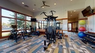 Located at Cascade Creek in Colorado Springs, Colorado, this single-level fitness center offers a comprehensive workout environment designed to support a wide range of exercise routines. The main workout floor is generously sized, featuring a multi-station cable machine, free weights, stability balls, and resistance equipment, all set beneath elevated ceilings with ceiling fans and recessed lighting. Mounted flat-screen TVs are positioned throughout for convenient viewing during workouts.

A dedicated spin studio accommodates multiple stationary cycling bikes arranged in rows, complete with a wall-mounted display screen and natural light filtering through windows with blinds. A separate TRX Training Zone provides a wall-mounted rack stocked with medicine balls, suspension trainers, and functional fitness accessories.

The cardio section is highlighted by floor-to-ceiling windows framed in rich wood trim, offering bright natural light and views of the surrounding Colorado landscape, including mature trees and greenery. Treadmills and elliptical machines are positioned to take full advantage of these outdoor views.

Throughout the facility, bold geometric patterned carpet tiles in blue, tan, and neutral tones add a modern aesthetic. Warm wood trim accents the walls, doorways, and window frames, giving the space a polished, welcoming character. A tiled entryway corridor with a water fountain provides a clean transition into the facility.