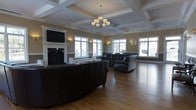 This refined single-floor lounge space showcases a sophisticated architectural design centered around a striking coffered ceiling with crisp white box beams and detailed crown molding. A decorative chandelier with frosted glass shades hangs as a focal point, complementing the room's classic aesthetic. The space is anchored by a traditional white-mantled fireplace framed by built-in cabinetry, above which a wall-mounted flat-screen television is positioned for comfortable viewing. Dark leather sofas and chairs are arranged throughout, creating multiple seating groupings that invite conversation and relaxation. Warm-toned hardwood flooring runs throughout, adding richness and continuity to the space. Large, multi-pane windows line the walls on multiple sides, flooding the interior with abundant natural light while offering views of the surrounding exterior. Wall sconces are strategically placed to provide soft ambient lighting. The neutral taupe wall color, accented by white wainscoting and trim, ties the design elements together cohesively. The overall ambiance is welcoming yet polished, balancing comfort and elegance in a generously proportioned communal setting.