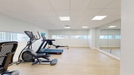 Located at 194 Tompkins Avenue in Beacon, NY, this fitness facility offers a clean, modern exercise space designed to support a well-rounded workout experience. The single-floor layout provides a generous, open area that feels airy and uncluttered, making it easy to move freely between equipment and open floor space.

The exercise room is finished with light-toned hardwood-style flooring that brightens the interior and complements the crisp white walls throughout. A drop ceiling with recessed panel lighting ensures consistent, even illumination across the entire space. A wall of floor-to-ceiling mirrors along one side expands the sense of space and supports both cardio and stretching routines.

A row of horizontal windows along the lower portion of the walls brings in natural light and offers views of the surrounding neighborhood. The room is equipped with a selection of cardio machines, including a treadmill, elliptical, rowing machine, recumbent bike, and a StairMaster, all arranged to allow ample room for use.

Additional amenities include a mounted flat-screen display, a dual water fountain with a bottle-filling station, a stainless steel waste receptacle, and yoga mat storage. A ceiling-mounted HVAC unit ensures comfortable climate control year-round. Colorful artwork displayed on the walls adds a welcoming, energetic touch to the overall ambiance.