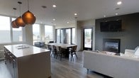 This single-floor space showcases a thoughtfully designed open-concept layout that seamlessly blends living, dining, and entertaining areas. The interior is finished with wide-plank hardwood-style flooring in a cool gray tone, complementing the clean, neutral walls and contemporary aesthetic throughout.

The dining area serves as a central focal point, anchored by a substantial rustic wood dining table surrounded by dark wood chairs — a striking contrast that adds warmth and character to the modern setting. Overhead, recessed lighting provides even, ambient illumination across the space.

The living area features a sleek sofa and is anchored by a wall-mounted flat-screen television above a modern gas fireplace set against a dark accent wall, creating a cozy yet sophisticated atmosphere.

Large floor-to-ceiling windows and a set of French doors flood the space with natural light and open directly onto an outdoor terrace, offering panoramic views of the surrounding cityscape and landscape. A secondary door provides additional outdoor access, reinforcing the strong connection between the interior and exterior living spaces.

A pendant light fixture with a warm copper tone adds a refined decorative touch, tying together the space's blend of rustic and contemporary design elements. Overall, the space exudes a bright, welcoming, and stylish ambiance ideal for both everyday living and entertaining.