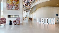 Located at 645 Division St in Nashville's evolving Wedgewood-Houston neighborhood, Albion Nashville presents a striking lobby and amenity floor defined by bold artistic vision and thoughtful architectural detail. The grand entrance greets residents with soaring double-height ceilings, polished concrete floors, and walls of floor-to-ceiling glass that flood the space with natural light while framing dynamic views of the Nashville skyline. A dramatic floral-patterned ceiling installation commands attention overhead, complemented by cascading globe pendant fixtures that add warmth and visual depth. The centerpiece reception desk features fluted wood paneling and is anchored beneath an illuminated archway, creating an elegant focal point. An expansive lounge area showcases rich hardwood flooring, vibrant watercolor murals stretching across full-length walls, and an eclectic mix of plush seating in mustard, plaid, and jewel tones set against magenta wainscoting. A separate seating nook offers an intimate retreat with additional mural art and varied chair styles. Corridor spaces display curated geometric artwork beneath deep blue-painted ceilings, adding a gallery-like quality to circulation areas. A dedicated package room with a vivid teal ceiling and lush greenery accents provides practical convenience. Throughout, the design balances artistic expression with functional comfort, reflecting Nashville's creative energy.