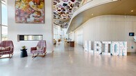 Located at 645 Division St in Nashville's evolving Wedgewood-Houston neighborhood, Albion Nashville presents a striking lobby and amenity floor defined by bold artistic vision and thoughtful architectural detail. The grand entrance greets residents with soaring double-height ceilings, polished concrete floors, and walls of floor-to-ceiling glass that flood the space with natural light while framing dynamic views of the Nashville skyline. A dramatic floral-patterned ceiling installation commands attention overhead, complemented by cascading globe pendant fixtures that add warmth and visual depth. The centerpiece reception desk features fluted wood paneling and is anchored beneath an illuminated archway, creating an elegant focal point. An expansive lounge area showcases rich hardwood flooring, vibrant watercolor murals stretching across full-length walls, and an eclectic mix of plush seating in mustard, plaid, and jewel tones set against magenta wainscoting. A separate seating nook offers an intimate retreat with additional mural art and varied chair styles. Corridor spaces display curated geometric artwork beneath deep blue-painted ceilings, adding a gallery-like quality to circulation areas. A dedicated package room with a vivid teal ceiling and lush greenery accents provides practical convenience. Throughout, the design balances artistic expression with functional comfort, reflecting Nashville's creative energy.