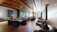 This thoughtfully designed single-floor communal lounge spans a generous open footprint, blending rustic warmth with contemporary sophistication. The architectural character is defined by striking exposed wood beam ceilings that soar overhead, complemented by rich wood-paneled walls and natural stone accent features that lend an inviting, lodge-inspired ambiance throughout the space.

The interior is organized into distinct yet fluid seating zones, each curated with stylish upholstered furniture in a palette of blues, grays, and warm earth tones. Plush swivel chairs, curved sofas, and accent armchairs are arranged around low coffee tables to encourage conversation and relaxation. A dedicated coffee bar area features a long wooden counter with bar-height seating, pendant lighting, and a full beverage station, serving as a functional social hub within the lounge.

Toward the entry, floor-to-ceiling glass doors and large windows flood the space with abundant natural light and provide views of the surrounding greenery outside. High pendant and track lighting fixtures throughout ensure the space remains well-lit during evening hours. Decorative art pieces, sculptural objects, and carefully chosen accessories add personality and visual interest across every zone. The wide-plank flooring and polished concrete surfaces ground the space with a tactile, organic quality that ties the entire design together seamlessly.