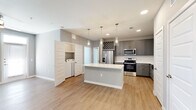 This well-appointed single-floor residence showcases a clean, contemporary design with a neutral gray and white color palette carried consistently throughout. The heart of the home is an expansive open-concept kitchen, anchored by a large central island with a crisp white quartz countertop and pendant lighting above. Gray shaker-style cabinetry lines the walls, complemented by a white tile backsplash, stainless steel appliances including a french door refrigerator, range, microwave, and dishwasher, and a farmhouse-style sink. A built-in wine rack adds a refined touch above the upper cabinets. Light wood-look flooring runs throughout the main living and kitchen areas, creating a warm, cohesive feel. A dedicated laundry nook is conveniently accessible from the main living space. The bedroom features plush carpeting, a ceiling fan, a natural light-filled window with blinds, and access to a walk-in closet. A secondary room with an exterior door and windows offers flexibility as a bonus room or dining area. The bathroom is elegantly finished with a dual-sink vanity, quartz countertops, gray cabinetry, a large mirror, and a tub-shower combination with marble-style tile surround. Recessed lighting and ceiling fans are featured throughout.