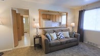This single-floor residence offers a practical and welcoming layout designed for comfortable everyday living. The living room serves as the heart of the home, featuring plush carpeted flooring and a warm, neutral color palette that creates an inviting atmosphere. A standout architectural detail is the wood-framed pass-through window that visually connects the living area to the kitchen, enhancing the sense of openness while maintaining a defined sense of space.

The kitchen is efficiently arranged with warm-toned wooden cabinetry providing ample storage, a white refrigerator, and a sink positioned beneath a window that allows natural light to filter in. The pass-through design not only adds a charming, rustic character to the space but also promotes easy interaction between the kitchen and living areas.

The living room is well-appointed, with enough space to comfortably accommodate a full sofa and side tables. Soft lighting from table lamps on either side of the seating area contributes to a cozy, relaxed ambiance. A hallway visible from the living room leads to additional areas of the home, suggesting a thoughtfully connected floor plan. Overall, the space balances functionality with a homey aesthetic, making it a comfortable and practical place to live.