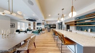 Located in Plymouth, Minnesota, this single-floor communal space offers a generously sized, open-concept layout that seamlessly integrates a kitchen, dining area, and living room. Light-toned hardwood flooring flows throughout, unifying the space and enhancing its airy, welcoming atmosphere.

The kitchen is a standout feature, boasting an expansive quartz-topped island with leather barstool seating and gold hardware accents. White cabinetry is complemented by a vibrant teal elongated tile backsplash and floating wood shelving. Pendant lights with leather-strap detailing add a distinctive, contemporary touch.

The dining area offers multiple seating configurations, including a large communal table, marble-topped bistro tables, and a cozy built-in banquette nook. The nook features deep teal accent walls with gold-tone horizontal trim details, plaid upholstered bench seating, and bold geometric artwork.

The living room showcases a floor-to-ceiling marble-surround fireplace flanked by a built-in wood bookshelf and a wall-mounted television. A sculptural black chandelier serves as an artistic focal point overhead. Plush seating in rich cognac leather and emerald green velvet is arranged to encourage conversation, while oversized windows flood the space with natural light and offer views of the surrounding landscape.
