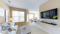 This single-floor residence offers an open-concept layout designed for comfortable, everyday living. The living area is spacious and welcoming, anchored by plush wall-to-wall carpeting and warmed by soft, neutral tones on the walls and ceiling. A large wall-mounted flat-screen TV is positioned above a sleek floating shelf, creating a natural focal point in the room. Generous windows fitted with horizontal blinds allow abundant natural light to fill the space, offering views of the surrounding greenery outside.

The living area flows seamlessly into a breakfast bar with bar stool seating, providing a casual dining option and a natural transition into the kitchen beyond. A pendant light fixture highlights the dining area tucked further in the background, where a small table and chairs are neatly arranged. The kitchen features counter space that opens to the bar, encouraging an interactive and social layout.

Throughout the space, the textured ceiling adds subtle architectural character, while the consistent warm beige palette creates a cohesive, calming ambiance. The single-floor design ensures easy accessibility and effortless movement between living, dining, and kitchen areas, making this a functional and inviting home.