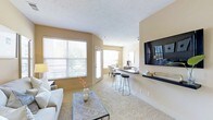 This single-floor residence offers an open-concept layout designed for comfortable, everyday living. The living area is spacious and welcoming, anchored by plush wall-to-wall carpeting and warmed by soft, neutral tones on the walls and ceiling. A large wall-mounted flat-screen TV is positioned above a sleek floating shelf, creating a natural focal point in the room. Generous windows fitted with horizontal blinds allow abundant natural light to fill the space, offering views of the surrounding greenery outside.

The living area flows seamlessly into a breakfast bar with bar stool seating, providing a casual dining option and a natural transition into the kitchen beyond. A pendant light fixture highlights the dining area tucked further in the background, where a small table and chairs are neatly arranged. The kitchen features counter space that opens to the bar, encouraging an interactive and social layout.

Throughout the space, the textured ceiling adds subtle architectural character, while the consistent warm beige palette creates a cohesive, calming ambiance. The single-floor design ensures easy accessibility and effortless movement between living, dining, and kitchen areas, making this a functional and inviting home.