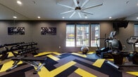 The Palm Living at Vintage Park Fitness Center is a thoughtfully designed workout facility that combines bold aesthetics with comprehensive fitness amenities. The space immediately captures attention with its striking chevron-patterned flooring in black, yellow, and brown — a dynamic design choice that energizes the environment and sets a motivating tone throughout.

The fitness center is illuminated by recessed ceiling lighting complemented by a large multi-blade ceiling fan, ensuring comfortable airflow during workouts. Full-length mirrors line portions of the walls, visually expanding the space and allowing members to monitor their form. Multiple windows along one wall invite an abundance of natural light and offer views of the surrounding outdoor area, including a glimpse of palm trees and a communal courtyard.

The facility is outfitted with a robust selection of equipment spanning both cardio and strength training. Cardio options include treadmills and an elliptical machine, while strength training is supported by a dumbbell rack, a weight bench, a multi-function cable machine, and a dedicated resistance training station. Stability balls and additional accessories further round out the offerings.

Dark gray walls lend a sleek, modern backdrop that contrasts sharply with the vibrant flooring, and motivational sports artwork adorns the walls. Wall-mounted televisions provide entertainment during workouts. The overall ambiance is energetic, clean, and purpose-built for an effective fitness experience.