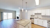 This single-floor, one-bedroom, one-bathroom residence offers a thoughtfully designed open-concept layout that maximizes its comfortable living space. The kitchen and living room flow seamlessly together, anchored by a central kitchen island with a neutral quartz-style countertop and elegant pendant lighting overhead. The kitchen is equipped with stainless steel appliances—including a gas range, built-in microwave, dishwasher, and French door refrigerator—complemented by crisp white shaker cabinetry and a classic subway tile backsplash. Light hardwood-style flooring extends throughout the main living areas, lending warmth and continuity to the space. The living room features a decorative fireplace with a detailed white mantel and crown molding, adding a refined architectural touch. Sliding glass doors open to a private balcony, allowing natural light to fill the room. The bedroom is carpeted for comfort, includes a ceiling fan, and provides access to a dedicated closet. The bathroom is outfitted with a full tub-shower combination, white tile surround, louvered closet doors, and hardwood-style flooring. Throughout the space, neutral tones and clean finishes create a bright, cohesive, and move-in-ready atmosphere.