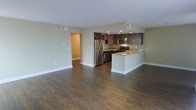 This single-floor space features an open-concept layout that integrates the kitchen and living area into one cohesive, functional space. The kitchen is outfitted with stainless steel appliances, including a refrigerator, microwave, and gas range, complemented by rich dark wood cabinetry and light-toned countertops. A peninsula-style counter extends into the main living area, providing additional workspace or casual seating while reinforcing the open flow between zones. Recessed lighting in the kitchen ensures ample illumination for food preparation. The living area is generously sized, offering a versatile space suitable for various furniture arrangements. Dark hardwood-style flooring runs throughout, providing a warm and grounding contrast to the light sage-green walls and bright white trim and ceiling. Visible HVAC vents are integrated discreetly into both the ceiling and floor. A hallway visible in the background suggests access to additional rooms beyond the main living space. The overall ambiance is clean, modern, and welcoming, with a neutral palette that lends itself well to a variety of interior design styles.