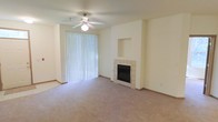 This single-floor space centers around a comfortable living area finished with neutral wall-to-wall carpeting and light-toned walls that create a bright, open atmosphere. A tile-surround fireplace serves as a focal point, complemented by a built-in niche above, ideal for displaying décor or a television. A ceiling fan with an integrated light fixture provides both air circulation and ambient lighting, enhancing the room's functionality. A large sliding glass door fitted with vertical blinds allows natural light to fill the space while offering the option for privacy, and likely provides access to an outdoor area. The clean, understated design throughout the living area offers a neutral canvas well-suited to a variety of furnishing styles. The overall layout is straightforward and practical, with an efficient use of space across the single level. The combination of the fireplace, natural light source, and ceiling fan makes the living area versatile and comfortable across seasons.