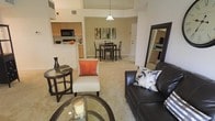 This single-story residence showcases an open-concept floor plan that integrates the living, dining, and kitchen areas into one cohesive, flowing space. The living area is carpeted and comfortably sized, furnished with a dark leather sofa, an accent chair, and a glass-topped coffee table, creating a relaxed yet polished setting. A large floor mirror along one wall adds depth and enhances the sense of space.

The dining area sits just beyond the living space, accommodating a dark wood dining table and matching chairs beneath a pendant light fixture. A colorful piece of wall art above the table serves as a focal point, adding a touch of personality to the area.

The kitchen is partially open to the main living space via a raised breakfast bar, featuring light wood cabinetry, dark appliances including a built-in microwave, and ample counter space. The layout allows for easy interaction between the kitchen and adjoining areas.

Notably high ceilings throughout amplify the open feel of the home, while neutral wall tones and consistent carpeting create a cohesive and inviting ambiance throughout the space.