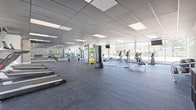 This fitness center offers a thoughtfully designed, single-floor space built to support a full range of workout routines. The open floor plan provides ample room to move freely between equipment zones, with durable dark rubber flooring throughout that is both functional and easy to maintain. A suspended dark tile ceiling fitted with recessed panel lighting creates a sleek, contemporary aesthetic while ensuring the space is well-lit.

One of the standout architectural features is the expansive floor-to-ceiling window wall that lines an entire side of the facility, flooding the interior with abundant natural light and offering open views of the surrounding outdoor greenery. This bright, airy quality gives the space an inviting and energizing atmosphere.

The fitness floor is comprehensively equipped with a dedicated cardio zone featuring treadmills, elliptical trainers, stationary bikes, and recumbent cycles. A strength training section includes a multi-functional cable machine, weight benches, free weights, and medicine balls. A central open area provides space for stretching and functional fitness exercises.

Mounted flat-screen televisions are positioned throughout the space for convenient viewing during workouts. A defining design element is the bold motivational word-art mural that spans a full wall, featuring phrases such as "Achieve," "Focus," "Believe," and "Inspire," reinforcing an atmosphere of encouragement and determination throughout the facility.