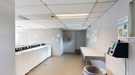 Located at 37140 S Orchard Cir in Westland, Michigan, this single-floor shared laundry facility offers a practical and efficiently organized space for communal use. The room is lined with rows of top-load washing machines and full-size dryers arranged along the perimeter and in central configurations, providing ample capacity for multiple users simultaneously. A drop ceiling with recessed fluorescent panel lighting ensures consistent, even illumination throughout the space. The neutral gray walls and light-toned vinyl tile flooring contribute to a clean, utilitarian aesthetic. Folding tables and countertop surfaces are positioned throughout the room, offering convenient workspace for sorting and folding laundry. Windows along one wall allow natural light to enter, adding brightness to the interior. A raised-step entryway with a door provides access, and a separate utility door is also present. Signage including a no-smoking notice is posted on the walls, indicating the facility's shared-use policies. The overall layout is open and unobstructed, allowing for easy movement between machines and work surfaces, making this a well-equipped and functional communal laundry facility.