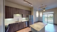 This one-bedroom, one-bathroom residence in Spring, TX is thoughtfully designed with modern finishes throughout. The kitchen serves as a central highlight, featuring dark wood cabinetry, a classic subway tile backsplash, and rich granite countertops that extend across a spacious central island — ideal for casual dining or meal prep. Stainless steel appliances, including a refrigerator, dishwasher, range, and microwave, complete the kitchen's contemporary aesthetic. Track lighting and elegant pendant lights illuminate the space, while wood-style flooring flows throughout the main living areas. A stacked washer and dryer unit is conveniently integrated within the kitchen area, adding practical functionality. The living area benefits from natural light and access to a private balcony, offering outdoor views and additional ventilation via a ceiling fan. The bedroom is carpeted for comfort, features its own ceiling fan, and is bathed in natural light through a large window with blinds. The bathroom is appointed with a granite-top vanity, dark wood cabinetry, and wood-style flooring, and connects directly to a walk-in closet with built-in shelving. The overall design balances modern style with everyday convenience throughout this single-level home.