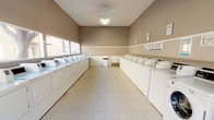 This shared laundry facility is a well-organized, single-floor space designed to accommodate the needs of a large residential community. The room is laid out with rows of white top-load washing machines and dryers lining both sides, providing ample capacity for multiple users at once. The light tile flooring is clean and easy to maintain, while the two-tone taupe and cream walls give the space a neutral, tidy appearance. A ceiling fan overhead aids in air circulation, complemented by bright overhead lighting that ensures the room is well-lit throughout. A row of windows along one wall allows natural light to filter in, adding to the overall brightness of the space. Informational signage is posted on the walls, offering guidance on machine usage and facility rules. A centrally located waste bin keeps the space orderly. The generous open floor area between the two rows of machines allows for comfortable movement and ease of use. Overall, this laundry facility is thoughtfully arranged to be functional, accessible, and efficient, serving as a practical communal amenity.