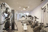 This fitness center is a fully outfitted single-floor facility designed to support a well-rounded workout routine. The space is furnished with a variety of professional-grade cardio equipment, including multiple Cybex treadmills, elliptical trainers, and stationary bicycles, all arranged to allow comfortable use and easy movement between machines. A multi-station strength training unit and additional free weight equipment provide comprehensive resistance training options, complemented by a padded workout bench for added versatility.

The room features neutral-toned textured walls and durable carpeted flooring suited for gym use. Recessed lighting throughout ensures the space is well-lit, while a ceiling fan provides added ventilation and comfort during workouts. Large windows and glass-paneled walls allow natural light to filter into the space, creating an open and airy atmosphere. Full-length mirrors line portions of the walls, enhancing the sense of space and allowing users to monitor their form during exercise.

Access to the fitness center is provided through glass-paned doors, with adjacent areas visible through the entryway. The layout is thoughtfully organized, offering a clear and functional flow that accommodates both individual and simultaneous use of the equipment.