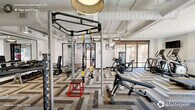 Located at 1005 West Main Street in Durham, NC, this fitness center offers a thoughtfully designed, spacious exercise environment spread across a single floor. The space is divided into two distinct zones that together provide a comprehensive workout experience.

The strength and cardio zone features a bold herringbone-patterned carpet tile floor and soaring ceilings with exposed ductwork, lending the area an industrial-modern aesthetic. It is outfitted with a functional training rig complete with TRX suspension straps and pull-up bars, weight benches, a cable machine, a leg press, and a dumbbell rack backed by floor-to-ceiling mirrors. Treadmills, an elliptical, a stair climber, and a rowing machine line the perimeter near large windows that invite natural light and outdoor views. Stylish drum pendant lights and ceiling fans complete the atmosphere.

The adjacent studio zone features smooth wood-look flooring and a cleaner, more open layout ideal for cycling and functional training. Smart spin bikes with integrated screens, a curated collection of kettlebells, medicine balls, and stability balls, along with wall-mounted TVs, make this area versatile. A dedicated shelving unit stocked with rolled towels adds a polished, amenity-rich touch. Glass partitions connect the two zones while maintaining visual separation, creating a cohesive yet multifunctional fitness facility.
