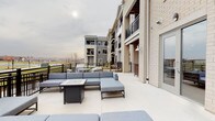 This single-floor community amenity space combines sophisticated interior design with inviting outdoor living. The main lounge area centers around a sleek, tiled linear fireplace framed by wood-beam ceiling details and a modern chandelier. Plush navy seating, soft area rugs, and warm stone-toned tile flooring create a refined yet comfortable atmosphere, while wall-mounted artwork and textured wallcoverings add visual depth. Glass sliding doors connect the interior seamlessly to a large outdoor terrace, flooding the space with natural light and offering unobstructed panoramic views of the surrounding landscape. The terrace itself is generously sized, furnished with sectional seating around a fire pit table and additional dining sets, making it ideal for gatherings. A dedicated dining and meeting area features multiple wooden tables paired with modern upholstered chairs, pendant lighting, and large windows that maintain a bright, open feel. Stone-look tile flooring flows consistently throughout, unifying the interior zones. The overall design balances contemporary aesthetics with warm, approachable finishes, resulting in a polished communal environment suited for both relaxed socializing and organized gatherings.
